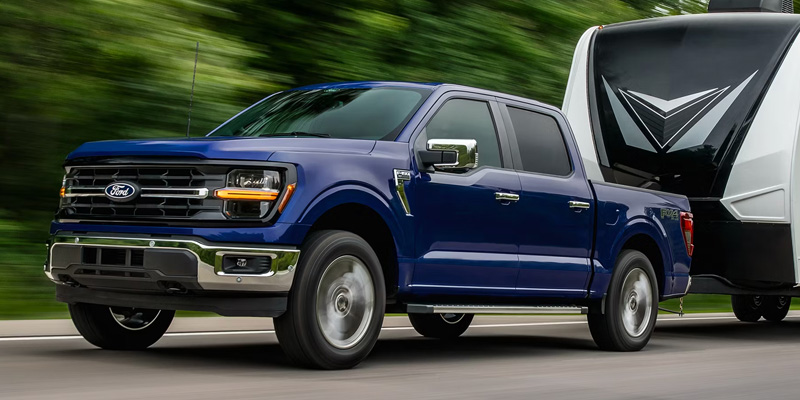 A blue Ford F-150 pickup truck towing a large white and black travel trailer down a highway.