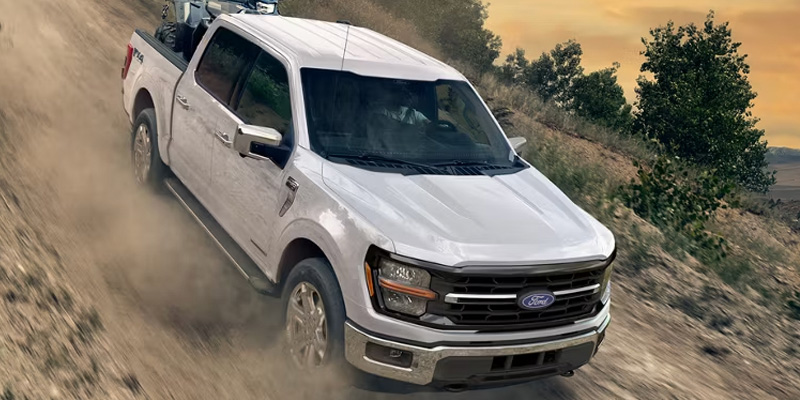 White Ford pickup truck driving off-road on a dirt trail, kicking up dust, with trees and hills in the background