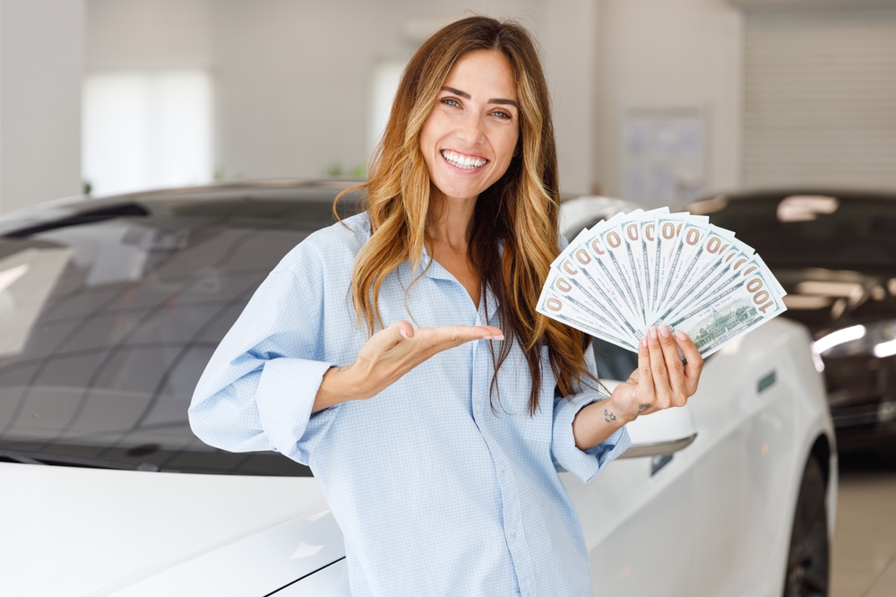 Woman smiling inside a car dealership showroom holding and displaying a fan of cash in her hand, standing in front of a white vehicle.