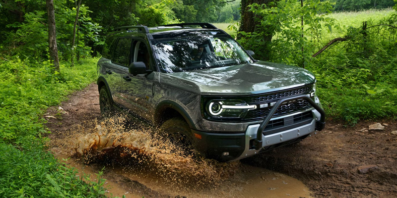 A Ford Bronco Sport SUV driving through a muddy forest trail, splashing water and dirt as it moves through off-road terrain.