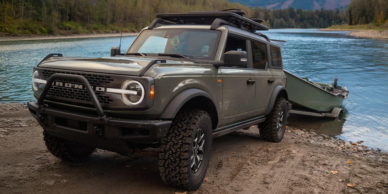 Front three-quarter view of a Ford Bronco-style SUV parked on a riverbank, towing a small boat with water and forested hills in the background.